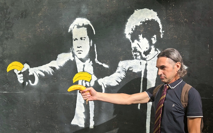 Man posing with Banksy mural of figures holding bananas at The Banksy Museum.