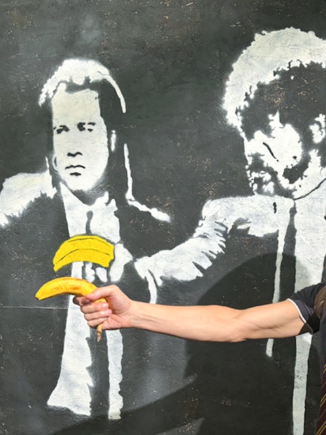 Man posing with Banksy mural of figures holding bananas at The Banksy Museum.