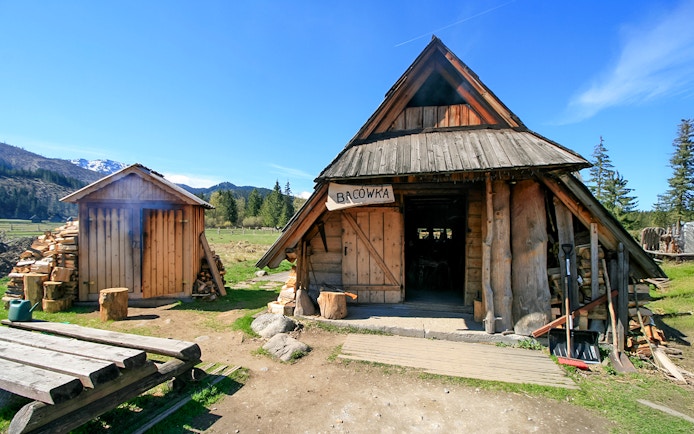 Bacowka Cheese Hut in a mountain meadow with wood stacks and clear sky.