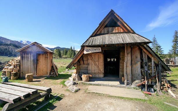 Bacowka Cheese Hut in a mountain meadow with wood stacks and clear sky.