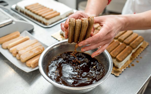 Hands dipping ladyfingers into coffee for tiramisù during cooking class.