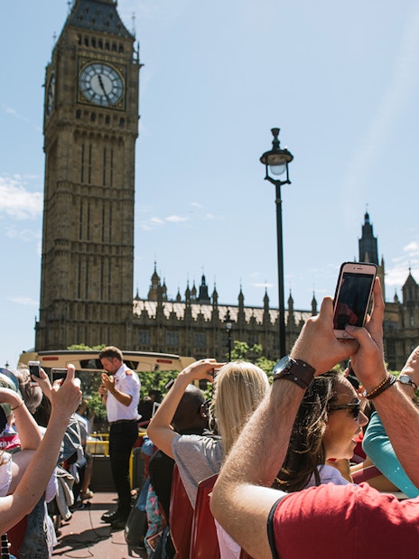 Tourists on a Big Bus near Big Ben in London, taking photos of the landmark.