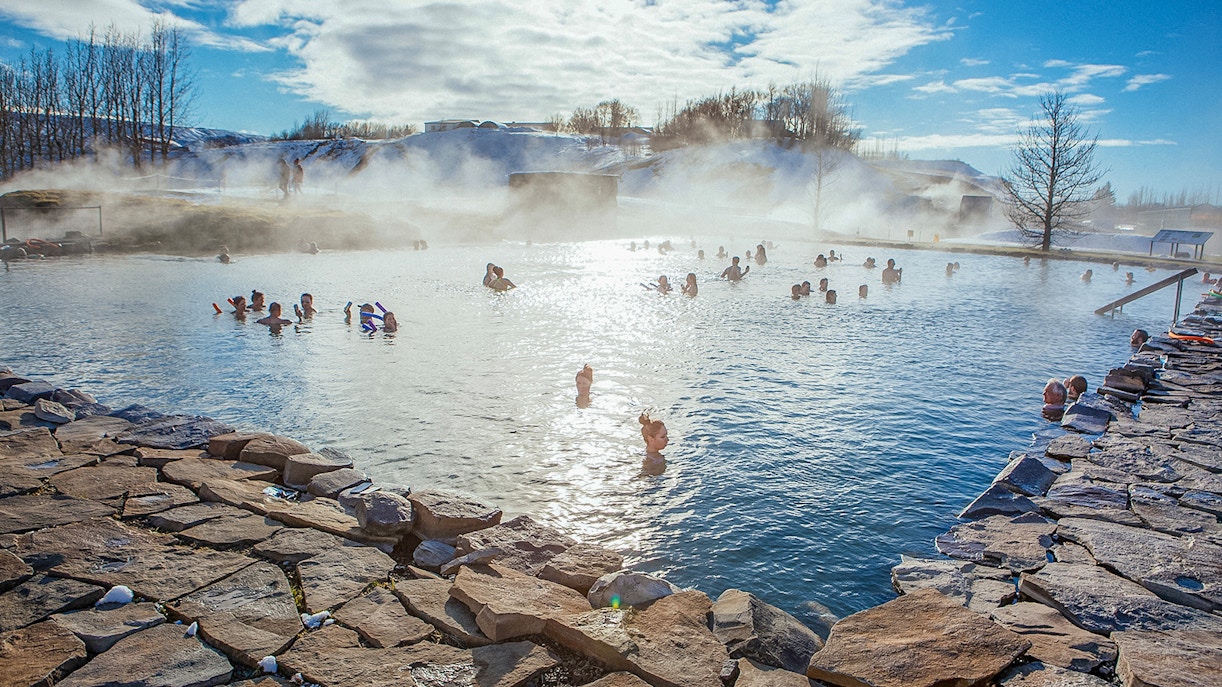 People enjoying a geothermal pool in Iceland with steam rising and snowy landscape.