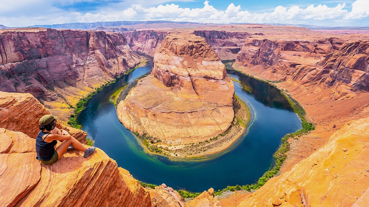 Person sitting on cliff edge overlooking Horseshoe Bend, Arizona, with river curving through canyon.