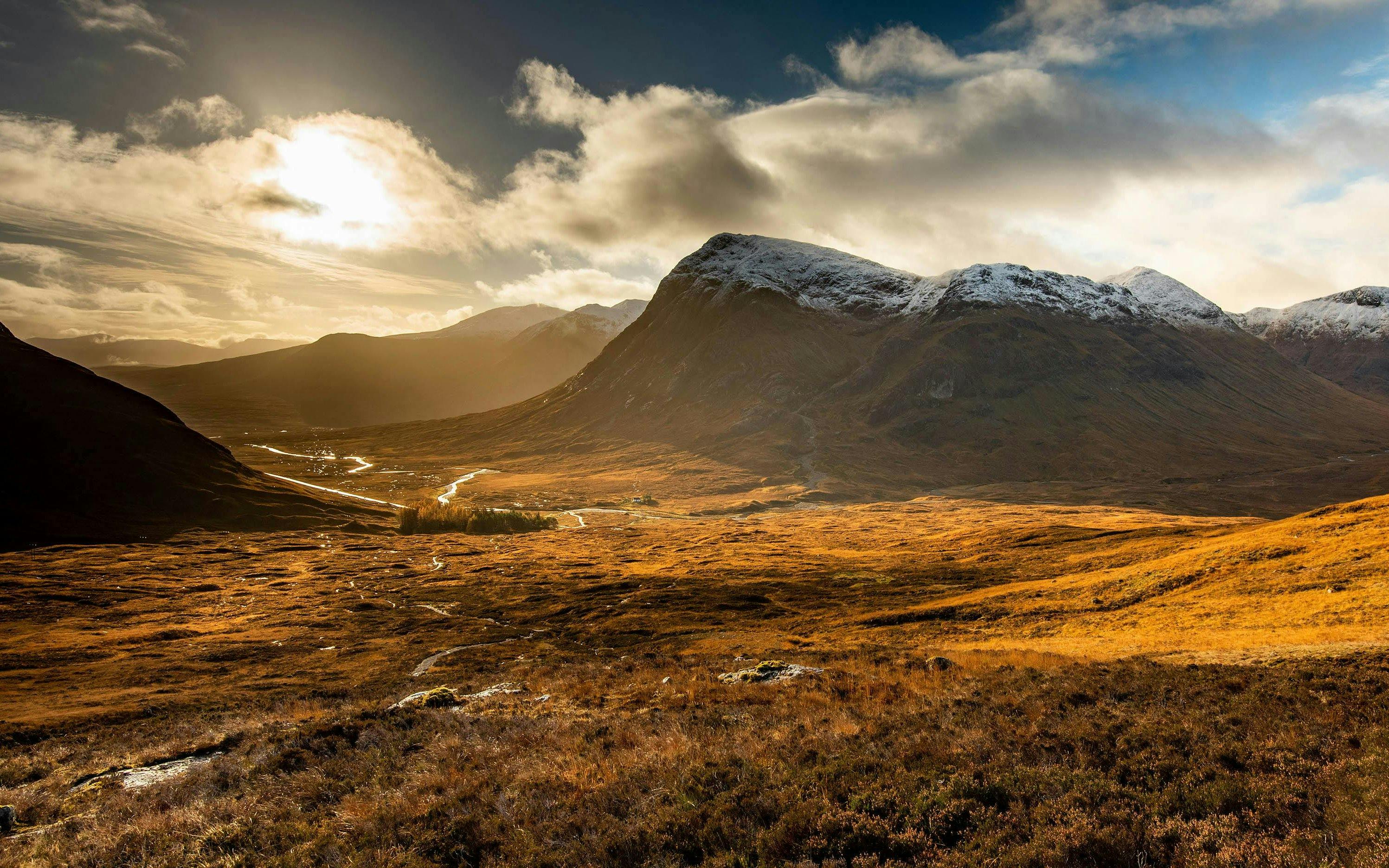 Sunlit valley and mountains in Glencoe, Scotland, with a winding river and cloudy sky.