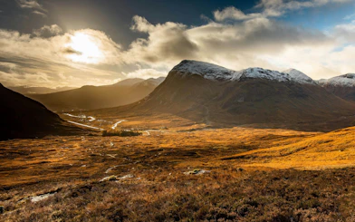 Sunlit valley and mountains in Glencoe, Scotland, with a winding river and cloudy sky.