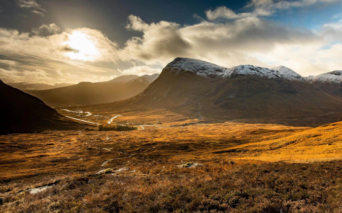 Sunlit valley and mountains in Glencoe, Scotland, with a winding river and cloudy sky.