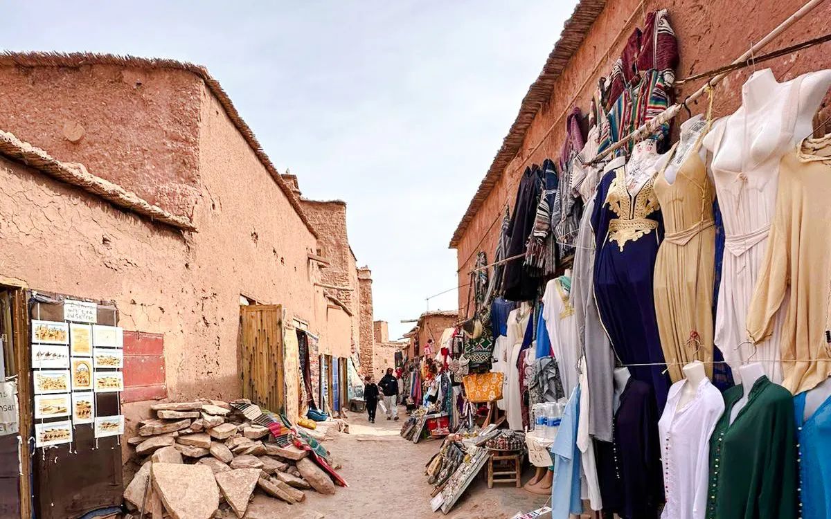 Market street in Merzouga with traditional clothing and goods displayed, part of 3 Days Desert Tour.