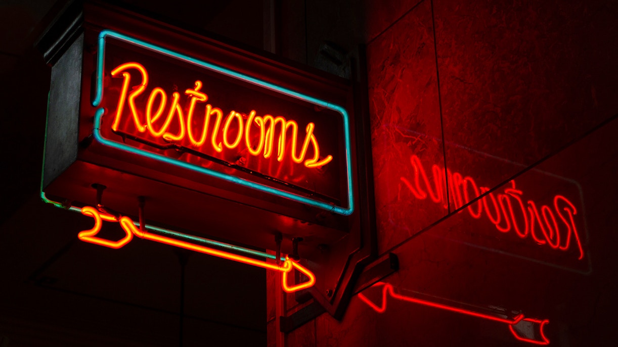 Neon "Restrooms" sign with reflection in a dimly lit urban setting.