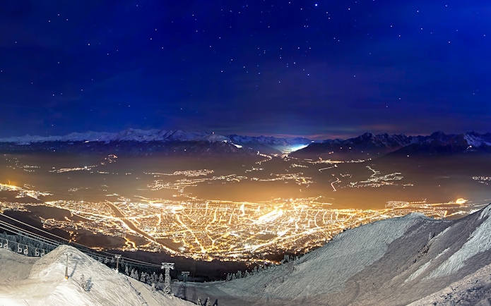 Nordkettenbahn view of Innsbruck city lights at night, with snowy mountains in the background.