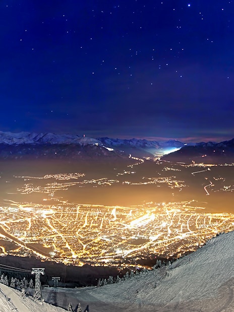 Nordkettenbahn view of Innsbruck city lights at night, with snowy mountains in the background.