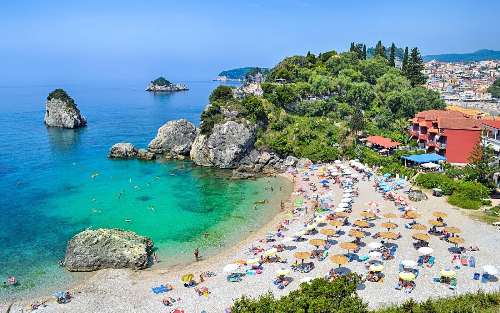 Parga beach in Corfu with sunbathers, umbrellas, and rocky coastline.