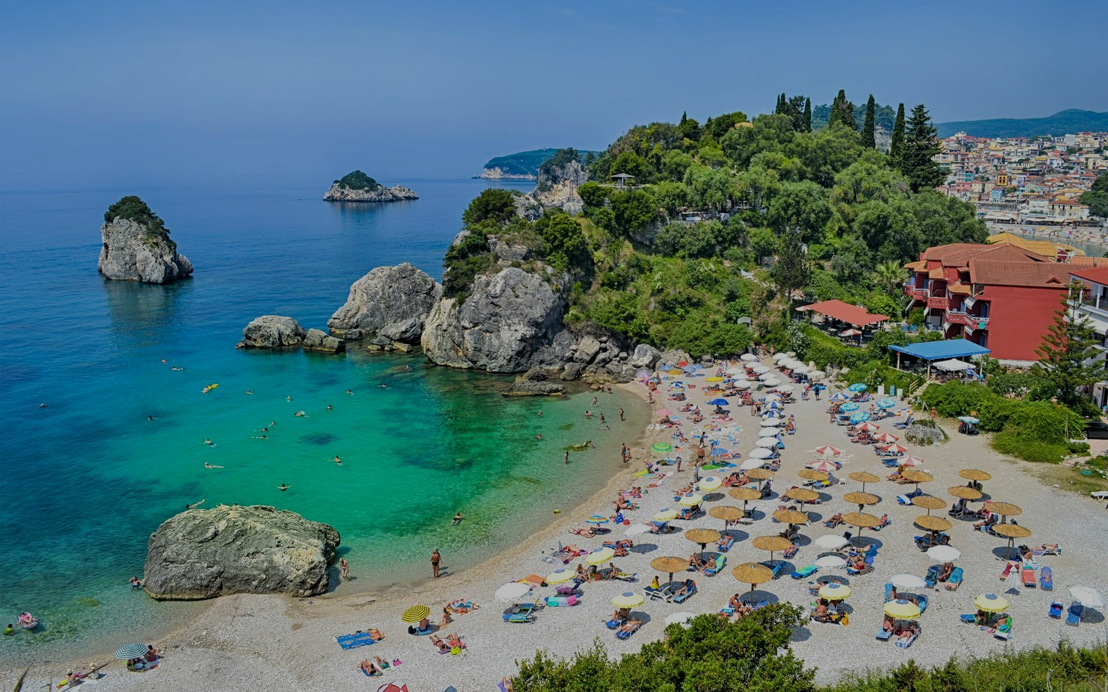 Parga beach in Corfu with sunbathers, umbrellas, and rocky coastline.