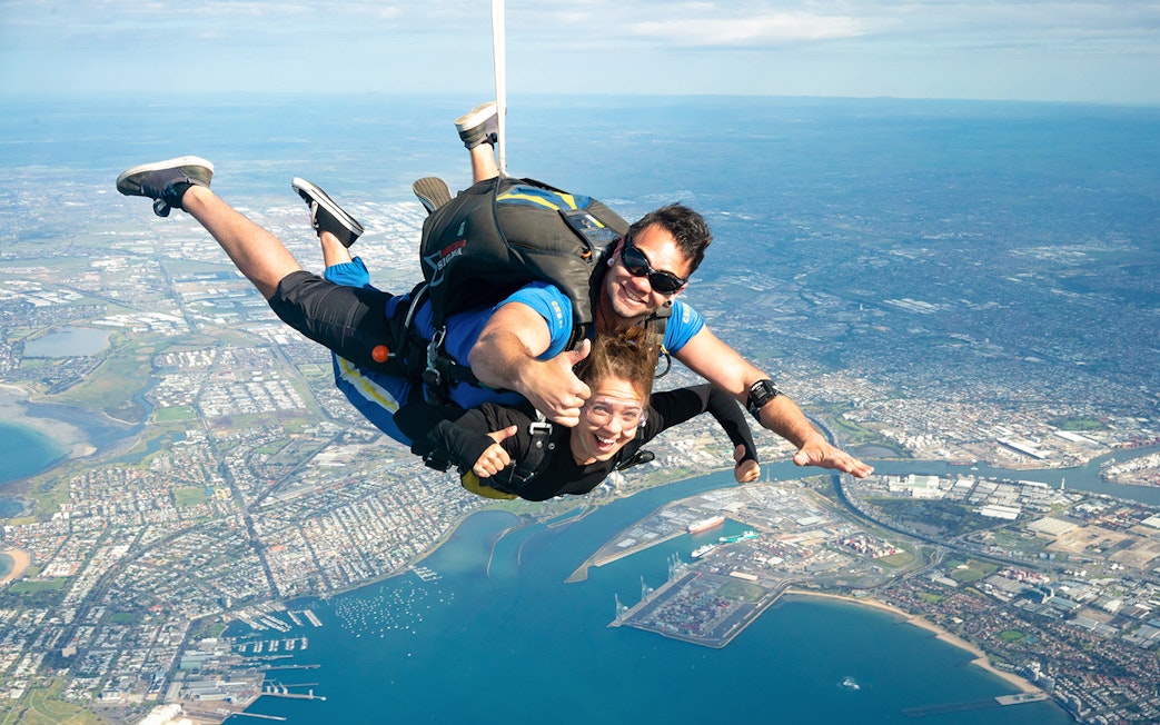 Tandem skydivers over St Kilda Beach, Melbourne with city and coastline below.