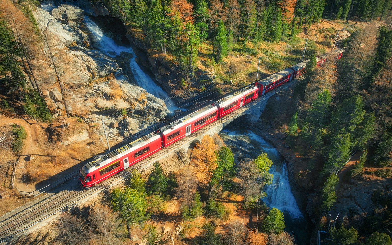 Aerial view of red passenger train traveling across a bridge over a waterfall, surrounded by colorful autumn forest in the swiss alps.