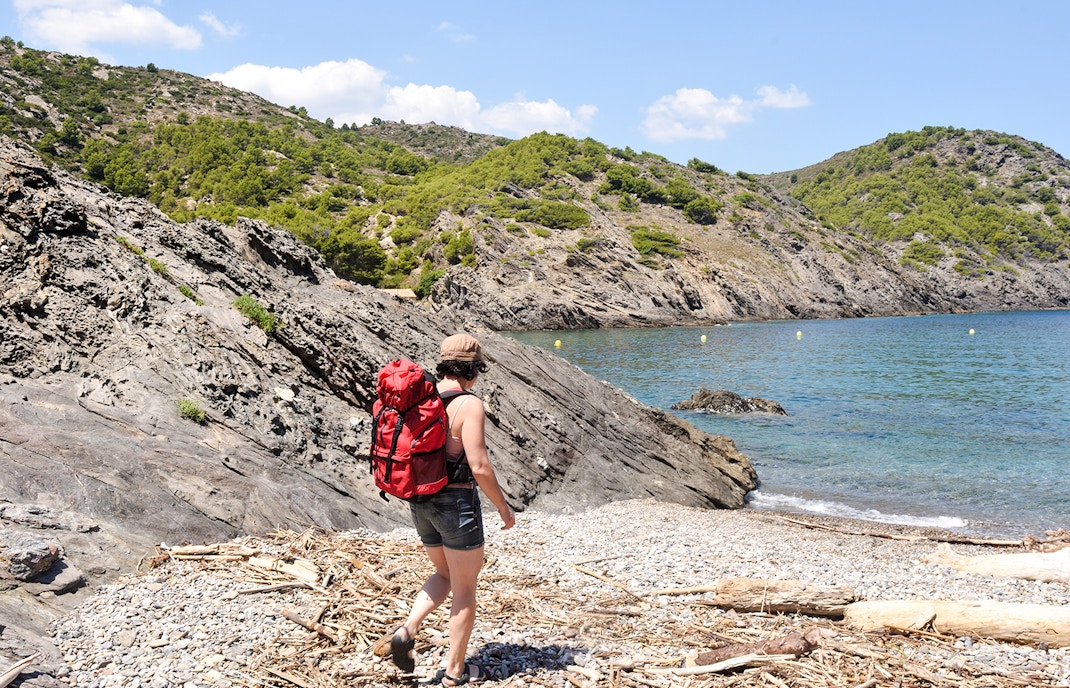 Hiker with red backpack on rocky Montjuïc trail by the sea.