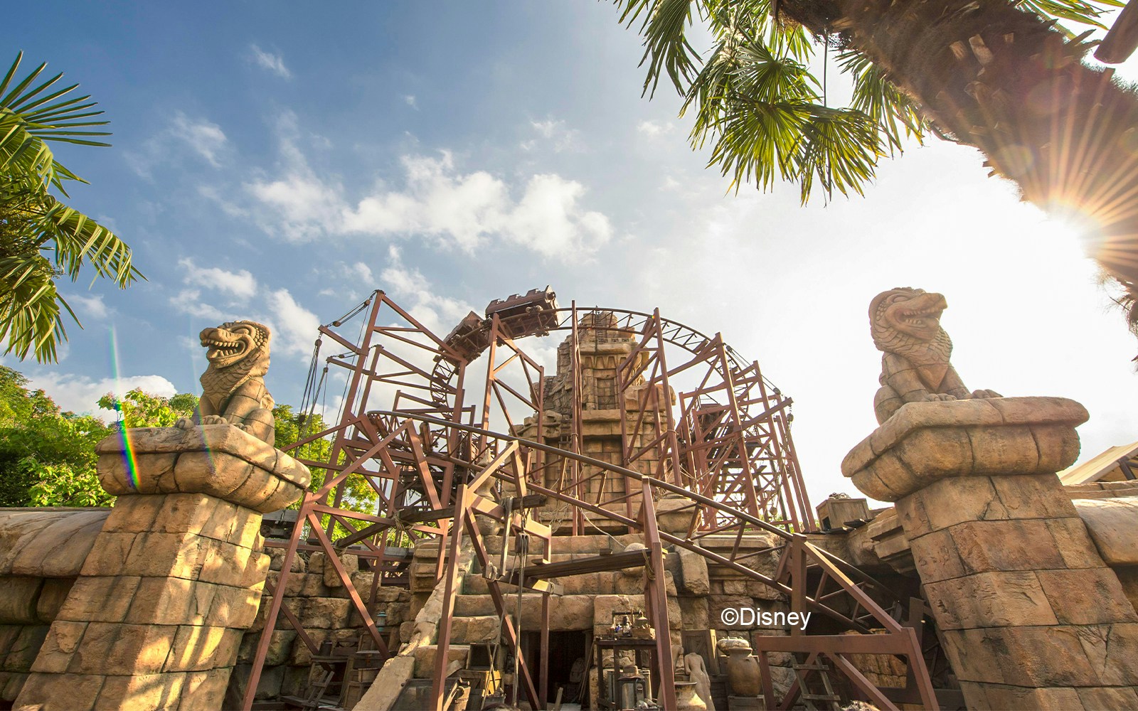 Roller coaster at Indiana Jones Adventure, Disneyland, with stone statues and palm trees.