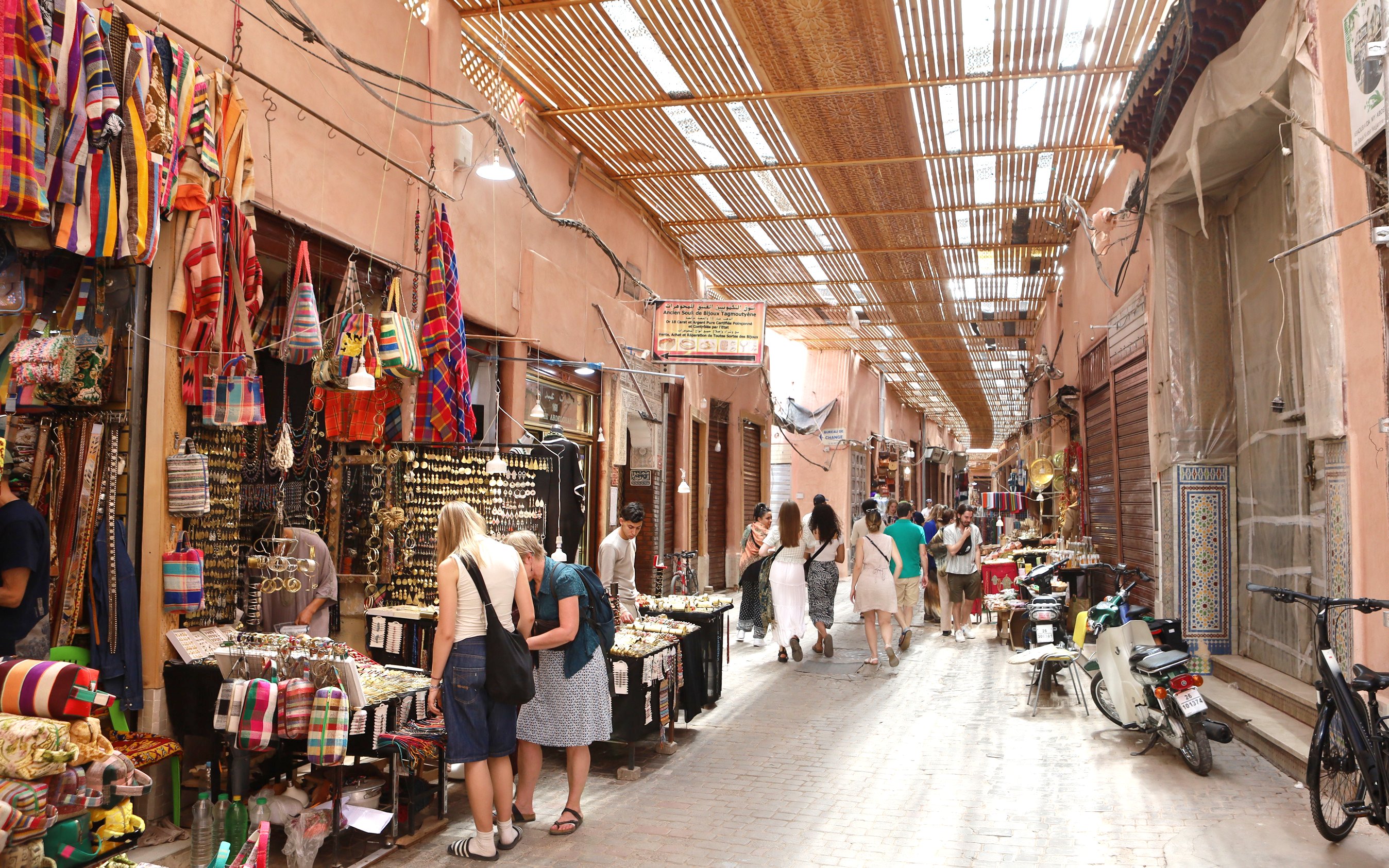Marrakech Medina market with tourists browsing colorful textiles and crafts.