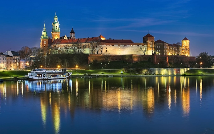Wawel Castle illuminated at night with a boat on the Vistula River in Krakow.