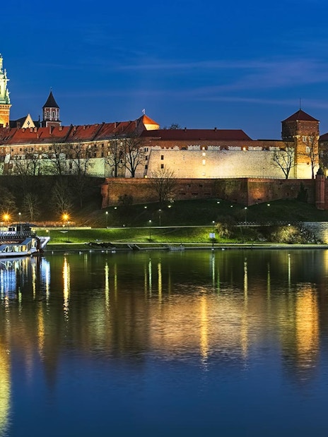 Wawel Castle illuminated at night with a boat on the Vistula River in Krakow.