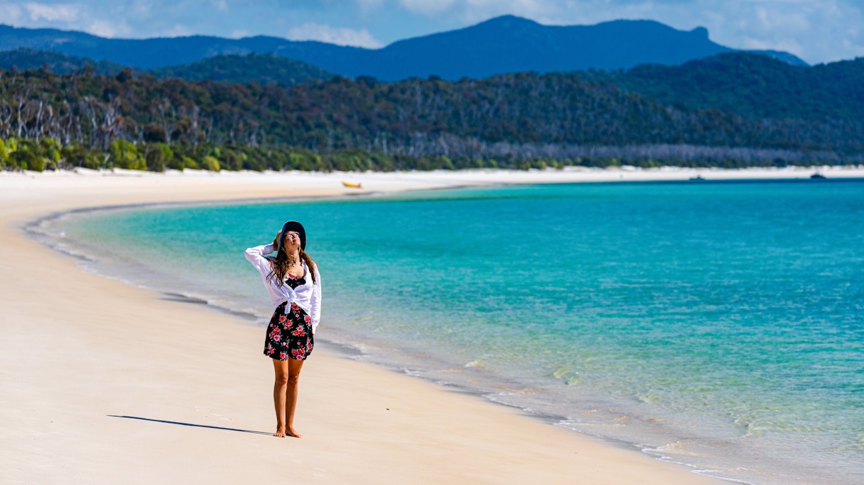 Person standing on Whitehaven Beach, Whitsunday Island, with turquoise water and forested hills.