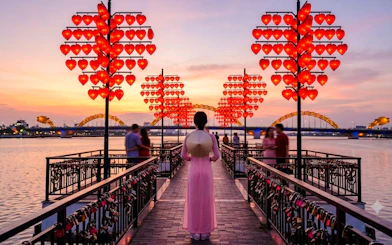Woman in traditional dress on Da Nang's Love Bridge at sunset with heart lanterns.