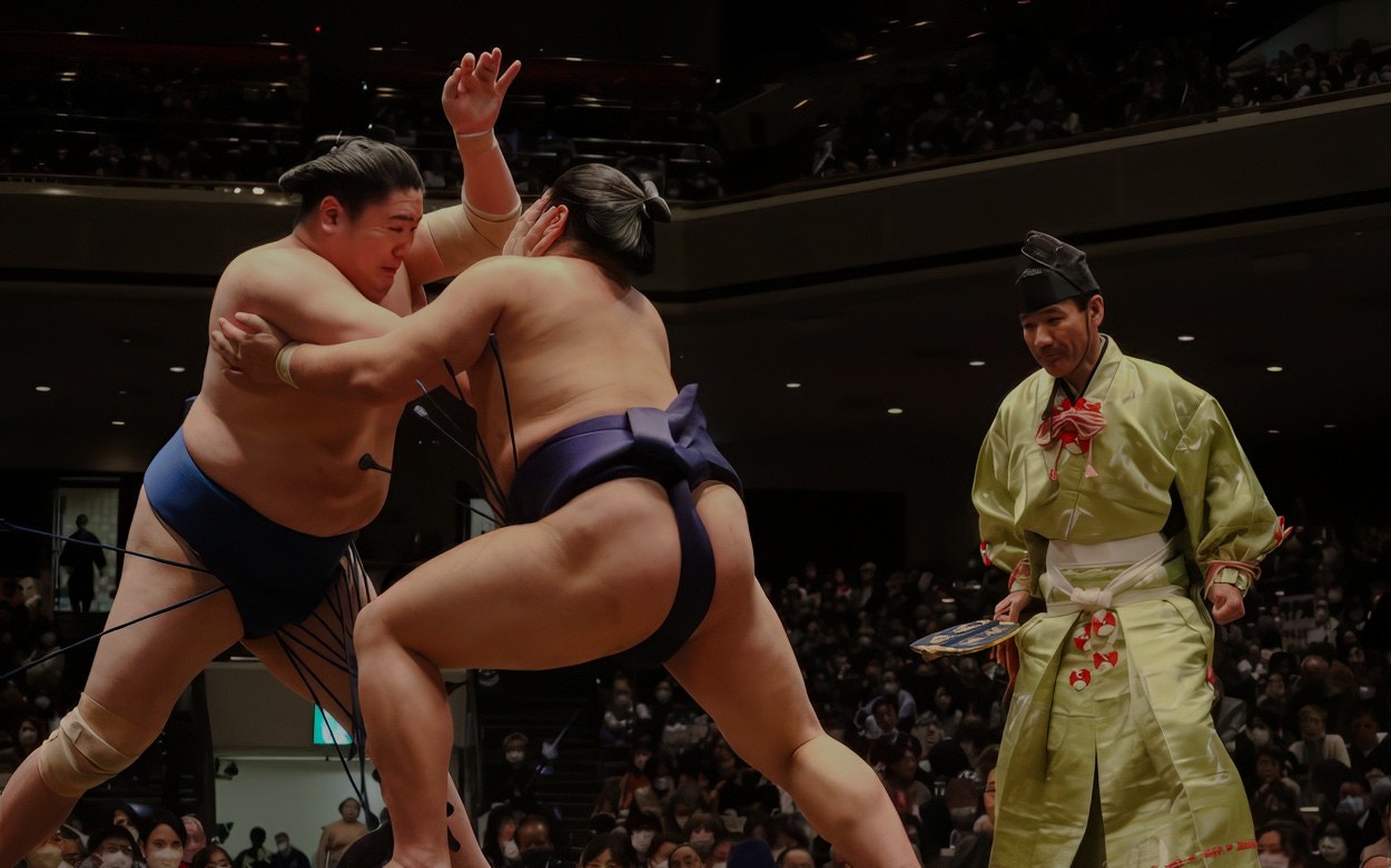 Sumo wrestlers competing in a Tokyo arena with a referee observing.