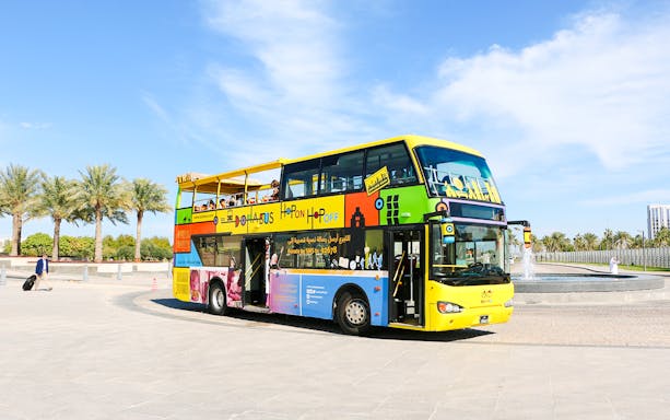 Colorful double-decker Doha Bus near palm trees and fountain.