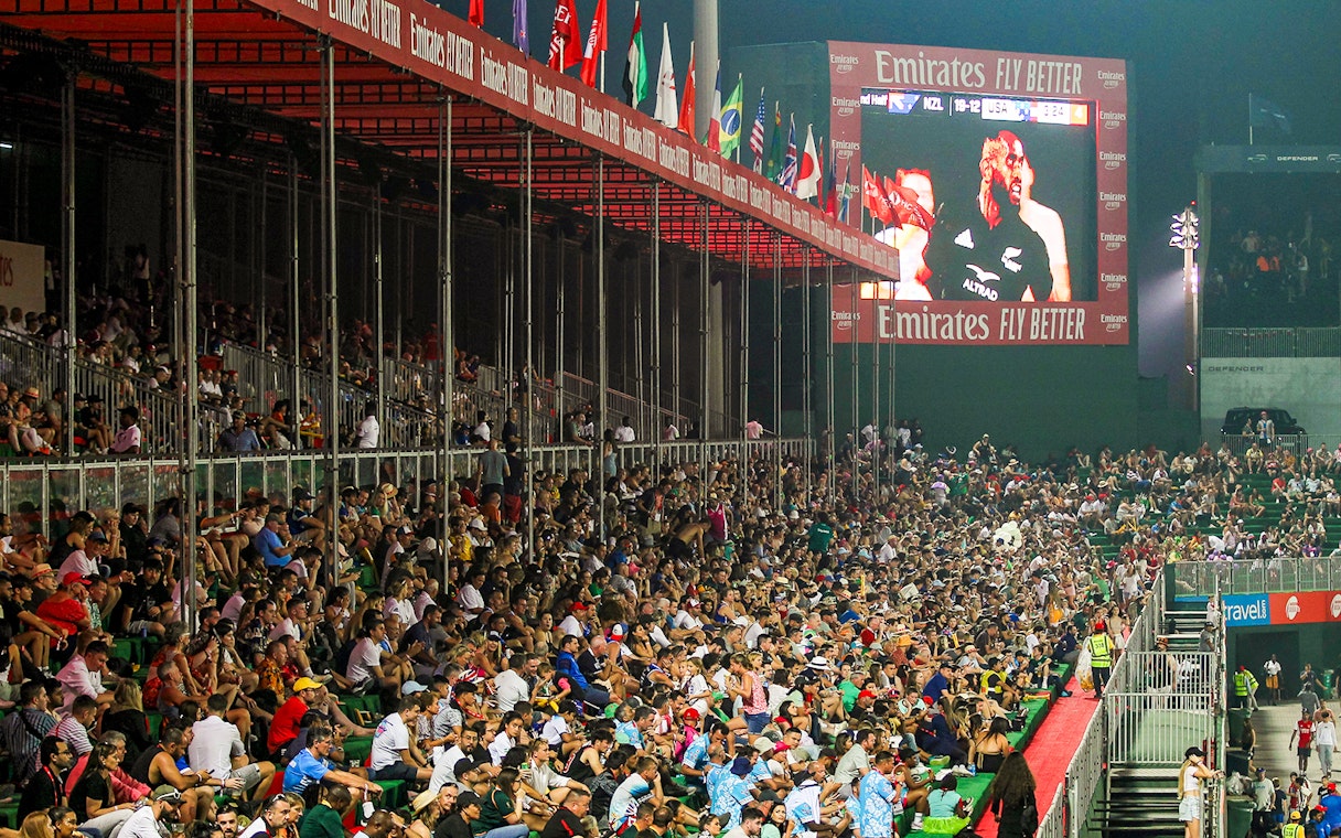 Crowd watching a rugby match at Emirates Dubai 7s stadium with flags and large screen.