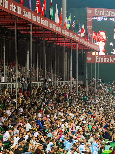 Crowd watching a rugby match at Emirates Dubai 7s stadium with flags and large screen.