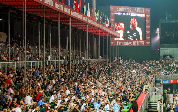 Crowd watching a rugby match at Emirates Dubai 7s stadium with flags and large screen.