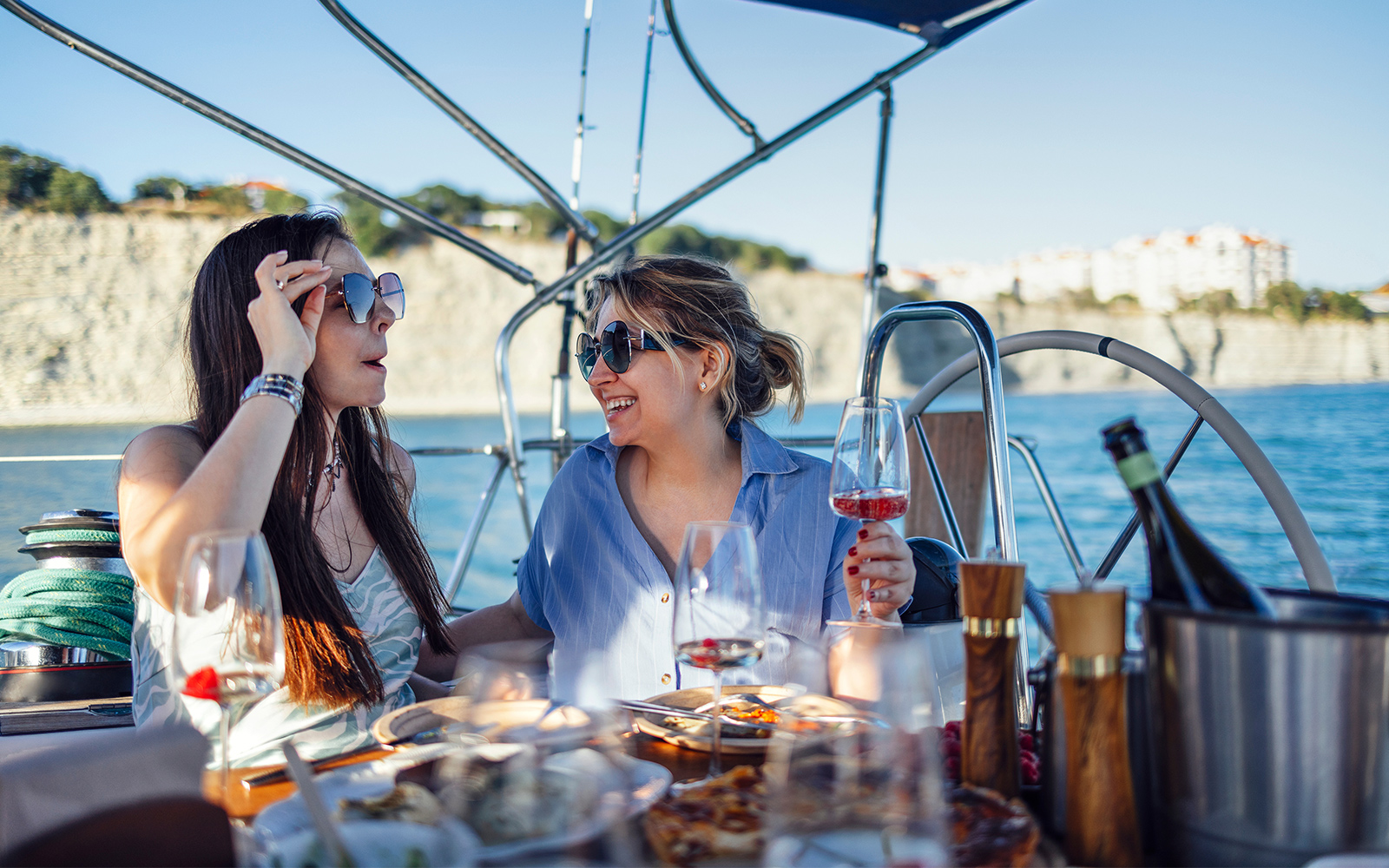 Two people enjoying a meal and drinks on a boat near a coastal cliff.