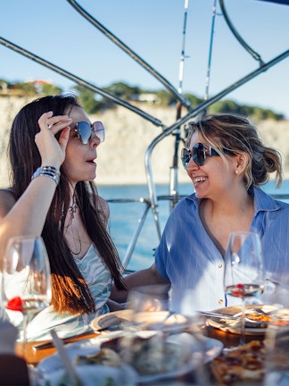 Two people enjoying a meal and drinks on a boat near a coastal cliff.