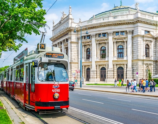 Red tram in front of Vienna's Burgtheater, Austria, with people walking nearby.