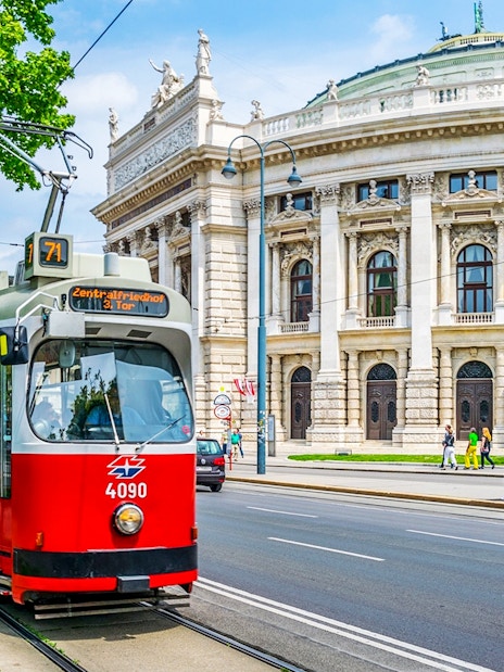 Red tram in front of Vienna's Burgtheater, Austria, with people walking nearby.