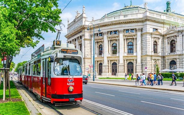Red tram in front of Vienna's Burgtheater, Austria, with people walking nearby.