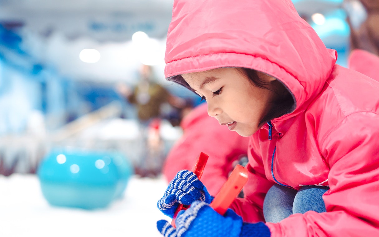 Child in red jacket playing at indoor ski park.