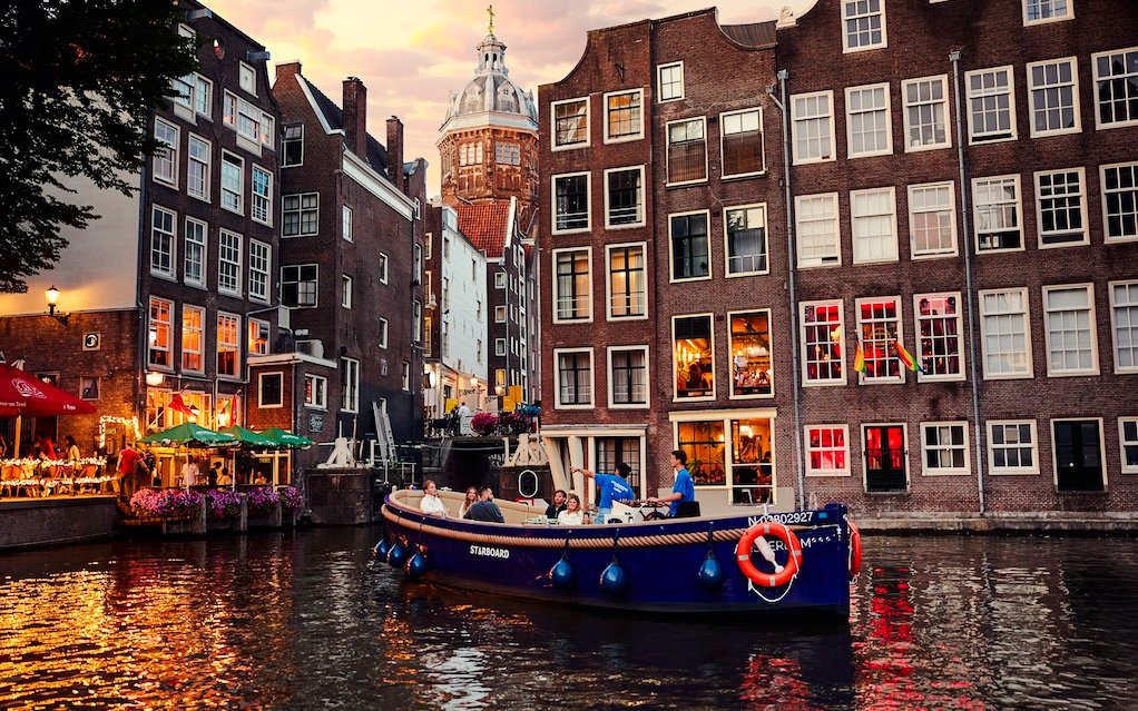 Covered canal cruise boat on Amsterdam canal with historic buildings at sunset.