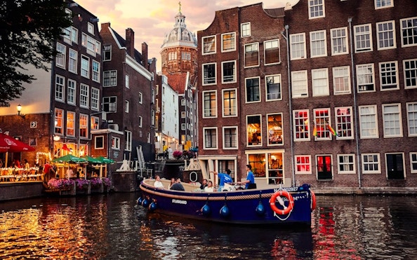Covered canal cruise boat on Amsterdam canal with historic buildings at sunset.