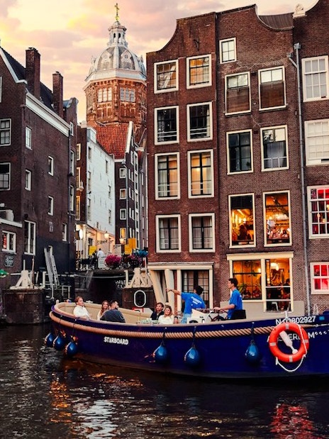 Covered canal cruise boat on Amsterdam canal with historic buildings at sunset.