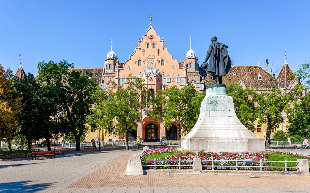 Town hall and statue in Kecskemét, Hungary, surrounded by trees and flowers.