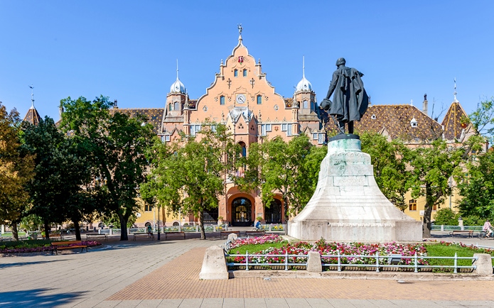 Town hall and statue in Kecskemét, Hungary, surrounded by trees and flowers.