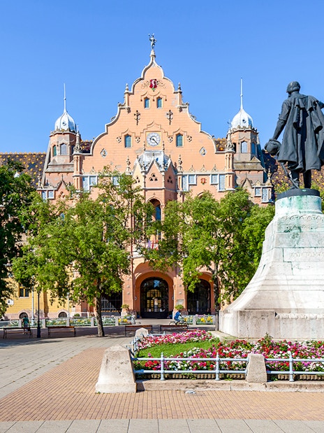 Town hall and statue in Kecskemét, Hungary, surrounded by trees and flowers.
