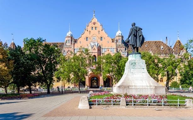 Town hall and statue in Kecskemét, Hungary, surrounded by trees and flowers.