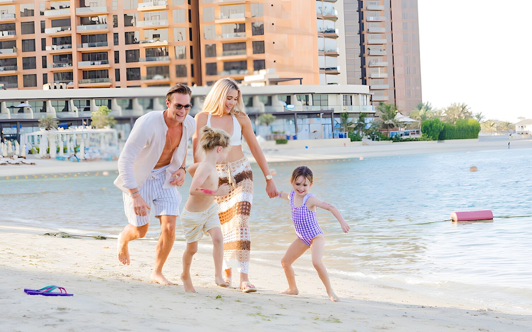 Family enjoying a day at Riva Beach Club, running along the sandy shore with buildings in the background.