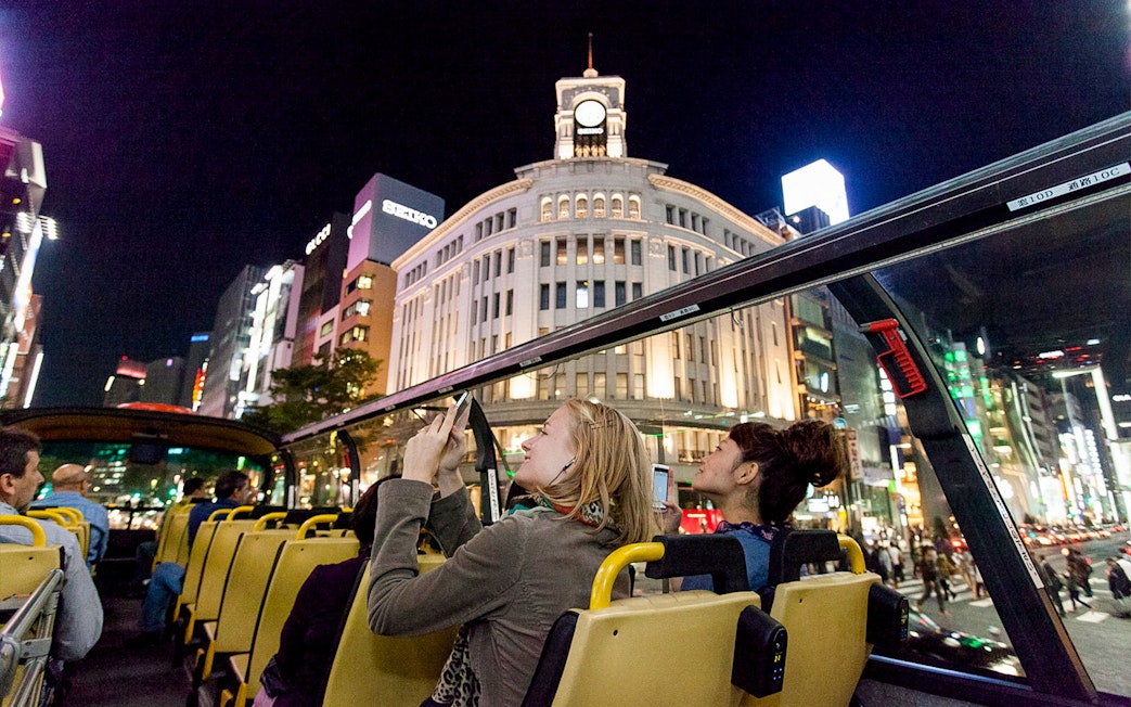 Open-top bus tour in Tokyo at night, passing by illuminated Ginza Wako building.