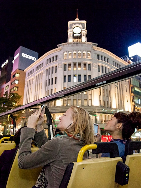 Open-top bus tour in Tokyo at night, passing by illuminated Ginza Wako building.