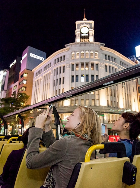 Open-top bus tour in Tokyo at night, passing by illuminated Ginza Wako building.