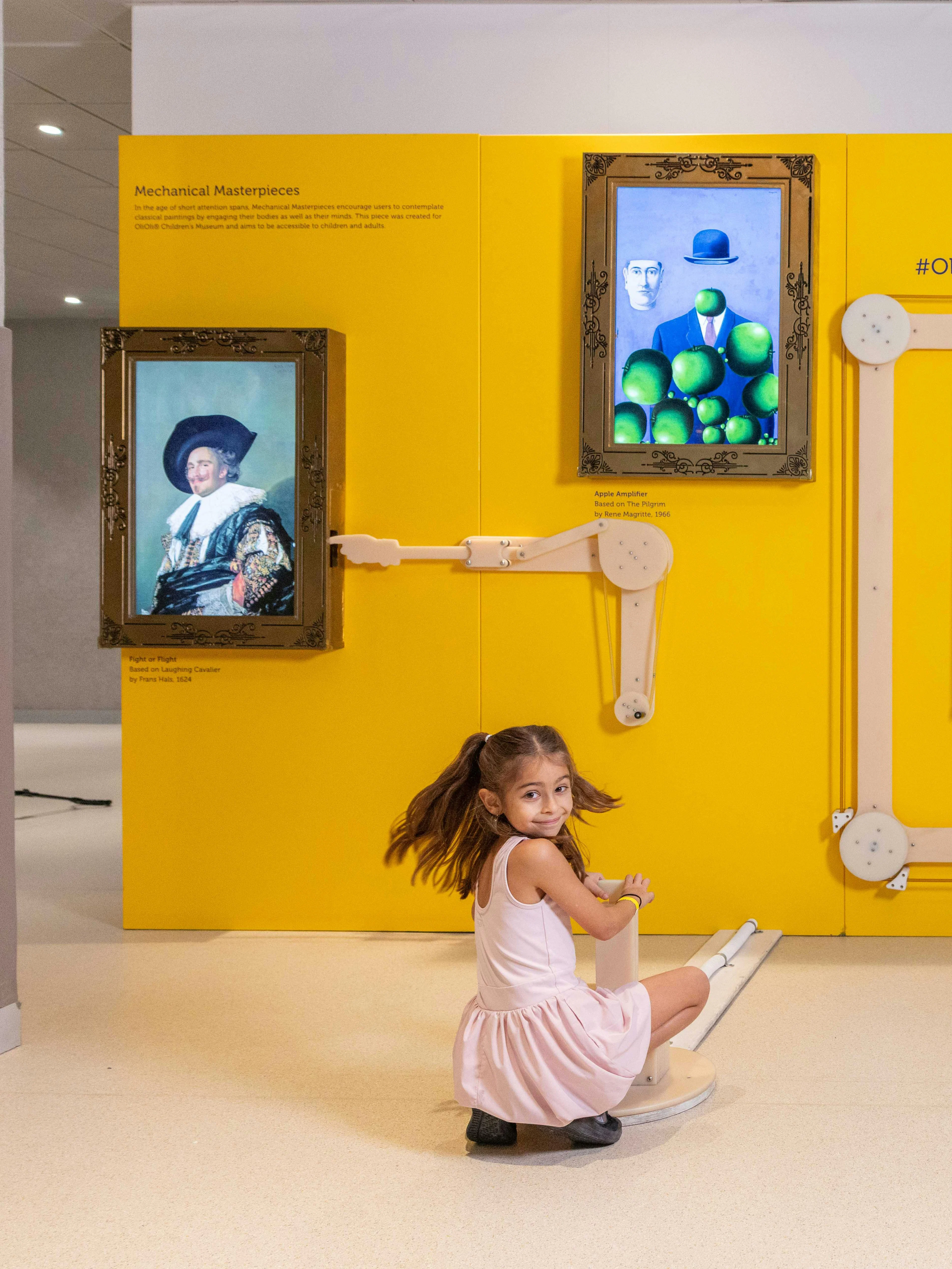Child interacting with art exhibit at OliOli Children's Museum, featuring famous paintings on a yellow wall.