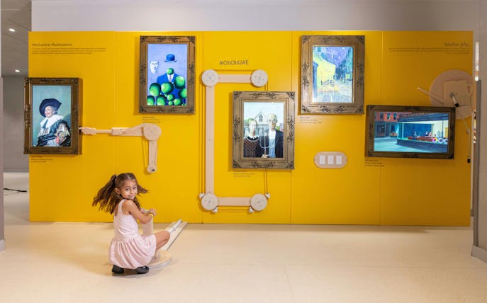 Child interacting with art exhibit at OliOli Children's Museum, featuring famous paintings on a yellow wall.